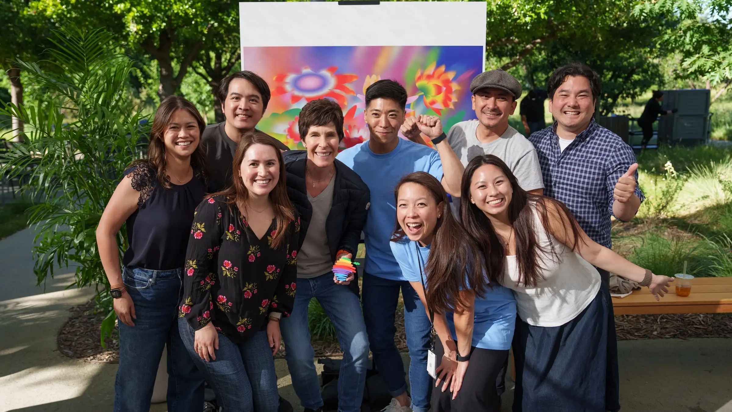 Nine members of the Asian Pacific @ Apple Silicon Valley chapter standing together and smiling outdoors at Apple Park, in front of a large colorful floral artwork on an easel