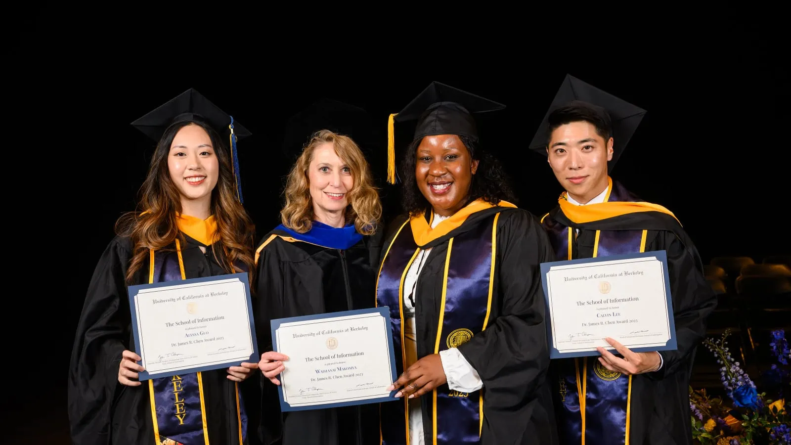 Four people in academic regalia on a commencement stage, three of them holding Dr. James R. Chen Award certificates from the School of Information